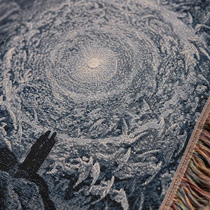 May include: A woven throw blanket featuring a dark blue and white intricate pattern of a swirling celestial vortex or sunburst. The fabric has a textured, tapestry-like weave with visible threads and a fringed edge displaying multicoloured fibres.