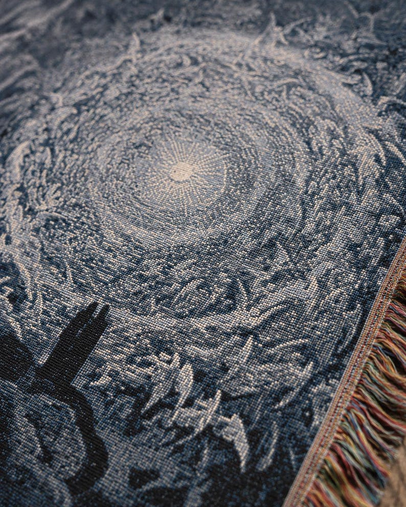 May include: A woven throw blanket featuring a dark blue and white intricate pattern of a swirling celestial vortex or sunburst. The fabric has a textured, tapestry-like weave with visible threads and a fringed edge displaying multicoloured fibres.