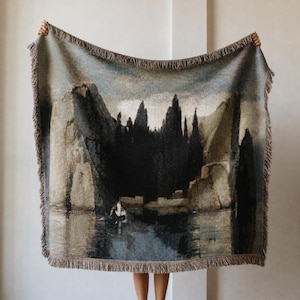 Puede incluir: Una manta tejida que presenta un paisaje oscuro y melanc&oacute;lico con acantilados imponentes, &aacute;rboles densos y un peque&ntilde;o bote sobre aguas tranquilas. La manta rectangular tiene bordes con flecos y una paleta de colores azul, gris y marr&oacute;n.