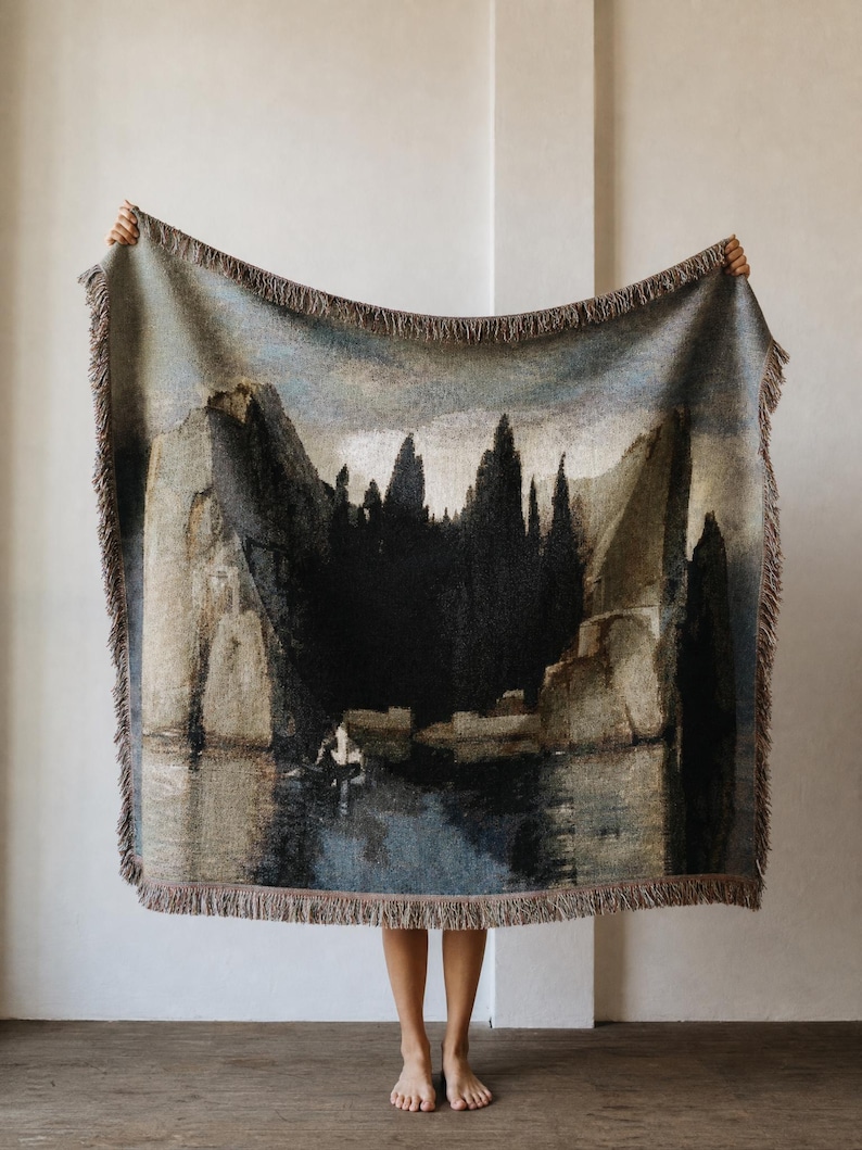 Puede incluir: Una manta tejida que presenta un paisaje oscuro y melanc&oacute;lico con acantilados imponentes, &aacute;rboles densos y un peque&ntilde;o bote sobre aguas tranquilas. La manta rectangular tiene bordes con flecos y una paleta de colores azul, gris y marr&oacute;n.