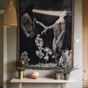 May include: A rectangular woven wall tapestry featuring a dark, monochromatic artistic scene of two figures, one with a bright beam of light emanating from their head, with fringed edges hanging against a light gray wall.