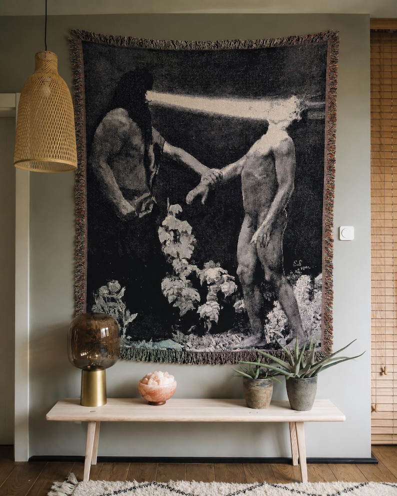 May include: A rectangular woven wall tapestry featuring a dark, monochromatic artistic scene of two figures, one with a bright beam of light emanating from their head, with fringed edges hanging against a light gray wall.