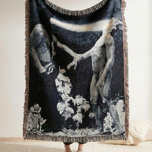 May include: A woven throw blanket featuring a dark blue background with a graphic illustration of two figures reaching toward each other, connected by a horizontal beam of light, finished with a fringed edge on all four sides.