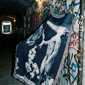 May include: A rectangular woven throw blanket featuring a dark blue background with a white graphic illustration of a figure standing near trees. The blanket has a fringed edge and displays the text THE NOHA STUDIO in the upper corner.