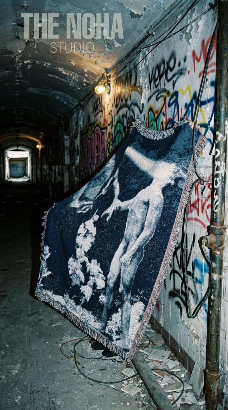 May include: A rectangular woven throw blanket featuring a dark blue background with a white graphic illustration of a figure standing near trees. The blanket has a fringed edge and displays the text THE NOHA STUDIO in the upper corner.