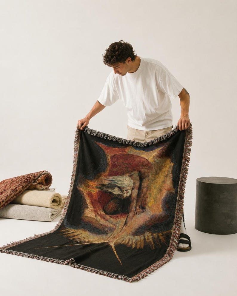 Puede incluir: Una manta tejida rectangular que presenta una reproducci&oacute;n de la pintura The Ancient of Days de William Blake. La obra muestra una figura que extiende un comp&aacute;s hacia abajo sobre un fondo oscuro y ardiente. La manta tiene bordes con flecos.