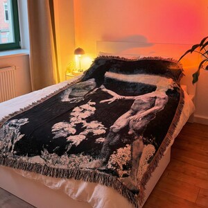 May include: A rectangular woven throw blanket featuring a high-contrast black and white graphic design of a human figure reaching out amidst textured, cloud-like shapes. The blanket has a decorative fringed edge along all four sides.