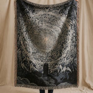May include: A rectangular woven throw blanket featuring a detailed monochromatic illustration of a celestial scene with a central radiating light source, dark swirling clouds, and two figures standing at the bottom, finished with fringed edges.