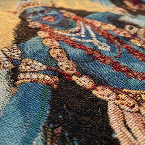 K&ouml;nnte beinhalten: Ein gewebter Wandbehang mit einer detaillierten Darstellung einer blauh&auml;utigen Figur, die mit geschichteten Halsketten und verziertem Schmuck geschm&uuml;ckt ist. Der Stoff zeigt eine reiche Farbpalette aus tiefem Blau, leuchtendem Gelb und Dunkelbraun.