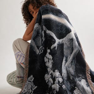 May include: A rectangular woven throw blanket featuring a dark, monochromatic artistic scene of two figures in a landscape. The blanket has a textured, tapestry-like appearance with fringed edges in muted tones of black, gray, and white.