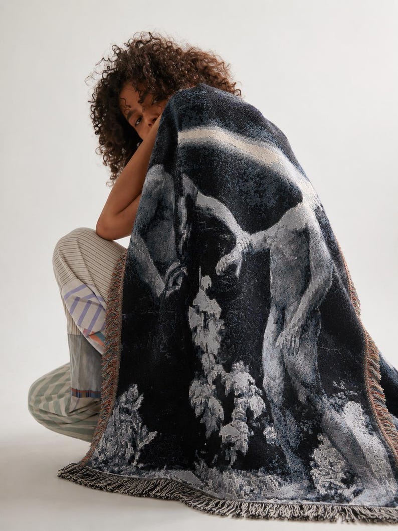 May include: A rectangular woven throw blanket featuring a dark, monochromatic artistic scene of two figures in a landscape. The blanket has a textured, tapestry-like appearance with fringed edges in muted tones of black, gray, and white.