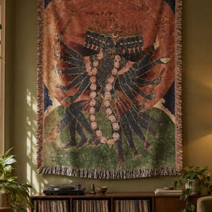 May include: A rectangular woven wall tapestry featuring a multi-armed dark-skinned figure with multiple heads, adorned with a large floral garland, set against a circular orange background, finished with a decorative fringed edge.