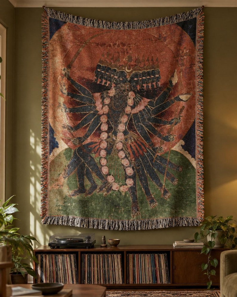 May include: A rectangular woven wall tapestry featuring a multi-armed dark-skinned figure with multiple heads, adorned with a large floral garland, set against a circular orange background, finished with a decorative fringed edge.