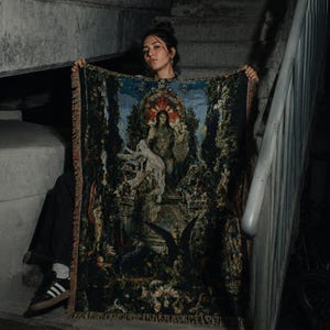Puede incluir: Una manta de tapiz tejida rectangular que presenta una ilustraci&oacute;n oscura y detallada de una figura central sentada en un trono rodeada de follaje exuberante, ruinas arquitect&oacute;nicas y criaturas aladas, con un borde de flecos.