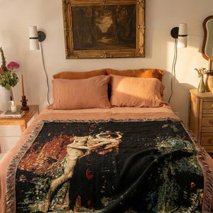 May include: A rectangular woven throw blanket featuring a dark black background with a detailed, colorful tapestry-style illustration of a figure holding a circular object amidst a landscape of trees and foliage, finished with fringed edges.