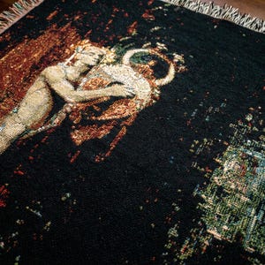 May include: A woven throw blanket featuring a dark black background with a detailed tapestry-style illustration of a figure embracing a large circular vessel. The edges of the blanket are finished with a decorative multi-colored fringe.