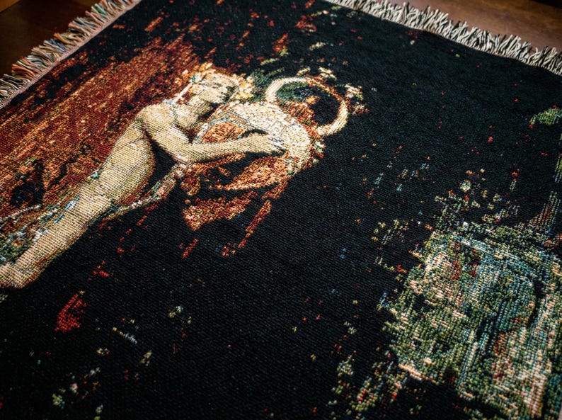 May include: A woven throw blanket featuring a dark black background with a detailed tapestry-style illustration of a figure embracing a large circular vessel. The edges of the blanket are finished with a decorative multi-colored fringe.