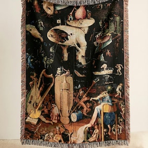 May include: Black woven throw blanket featuring a detailed print of a Hieronymus Bosch painting. The artwork depicts a surreal scene with figures and fantastical elements. The blanket has a fringed edge.