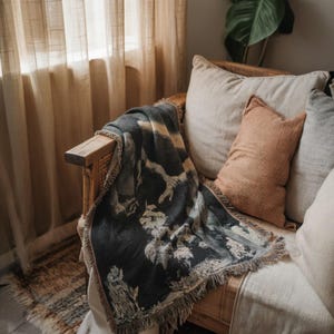 May include: A woven throw blanket featuring a dark charcoal and cream abstract pattern with fringed edges, draped over a wooden chair with a light beige cushion and a smaller textured terracotta-colored rectangular pillow.