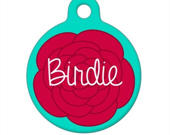 May include: A teal pet tag with a red rose design and the name "Birdie" in white script. The tag is round with a hole at the top for attachment to a collar. The design is simple and colorful.