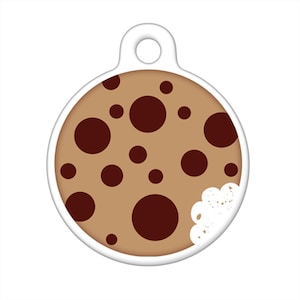 May include: A round, white dog tag with a brown and white cookie design. The cookie has chocolate chips and a bite taken out of it.