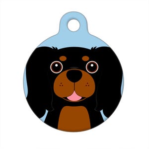 May include: A round dog tag with a cartoon image of a black and brown Cavalier King Charles Spaniel dog with big eyes and a happy expression. The background is light blue.
