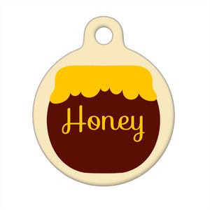 Op de afbeelding: Een ronde, crèmekleurige huisdiertag met een bruine honingpot-illustratie en het woord "Honey" in gele letters.