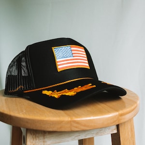 May include: Black trucker hat featuring an embroidered American flag patch. The hat has a mesh back and a curved brim. Gold embroidery accents the brim. The hat is sitting on a wooden stool.