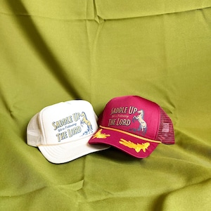 May include: Two trucker hats with mesh backs. One is white with a beige brim, the other is maroon. Both have the text "SADDLE UP We're Following THE LORD" and a horse graphic.