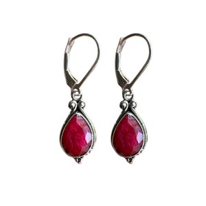 May include: A pair of silver-toned leverback earrings, each featuring a faceted, teardrop-shaped, deep red gemstone. The gemstones are set in a decorative bezel setting, adding a touch of elegance to the jewelry.