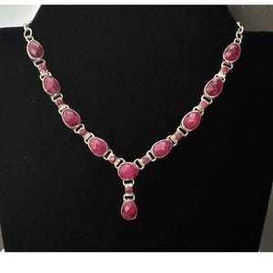 May include: A silver necklace featuring oval-shaped, deep pink gemstones. The necklace has a pendant with a teardrop-shaped gemstone. The gemstones are set in a silver metal chain.