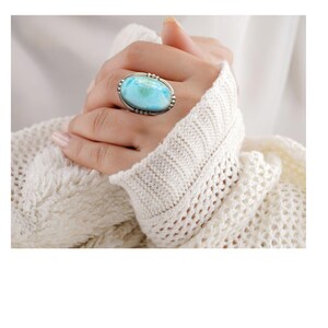May include: A silver ring featuring a large, oval-shaped, light blue gemstone. The ring has a detailed silver setting with small beaded accents around the gemstone. The ring is displayed on a hand, with a cream-colored knitted sweater in the background.