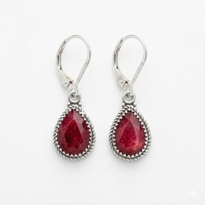 May include: A pair of silver leverback earrings with teardrop-shaped, faceted, deep red gemstones. The gemstones are framed by a twisted rope design. The earrings are displayed against a white background.