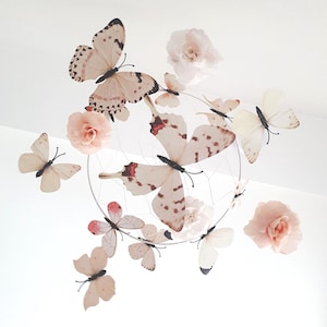 Puede incluir: Móvil decorativo con mariposas y flores. Las mariposas tienen alas color crema con manchas marrones y detalles rojos. Las flores son de color rosa claro. El móvil está suspendido de un aro plateado.