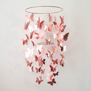 Könnte beinhalten: Ein roségoldenes Schmetterlingsmobile mit zahlreichen Schmetterlingen, die an zwei kreisförmigen Reifen aufgehängt sind. Die unterschiedlich großen Schmetterlinge sind kaskadenartig angeordnet und bieten ein elegantes Dekorationselement.