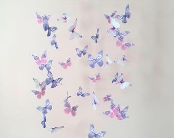 Könnte beinhalten: Ein Mobile mit zahlreichen zarten Schmetterlingen in Blau-, Rosa- und Weißtönen. Die Schmetterlinge scheinen an einem weißen, kreisförmigen Ring aufgehängt zu sein, was eine skurrile und luftige Ästhetik erzeugt. Die Schmetterlinge variieren in der Größe und sind so angeordnet, dass sie den Eindruck von Flug erwecken.
