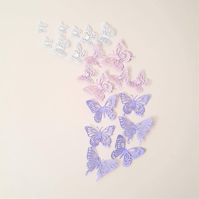 May include: A collection of decorative butterfly wall decals in various sizes and colors. The butterflies range from white to shades of pink and purple, creating a gradient effect. The butterflies are arranged in a flowing, upward pattern.
