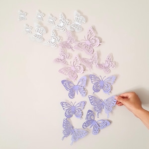 May include: A collection of paper butterflies in various shades of white, lavender, and purple are arranged on a light beige wall. The butterflies are in different sizes and have intricate cut-out designs, creating a whimsical and decorative display.
