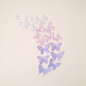 May include: A collection of paper butterflies in various sizes and shades of white, pink, and purple. The butterflies are arranged in a cascading pattern, creating a decorative wall art piece. The butterflies have intricate cut-out designs.