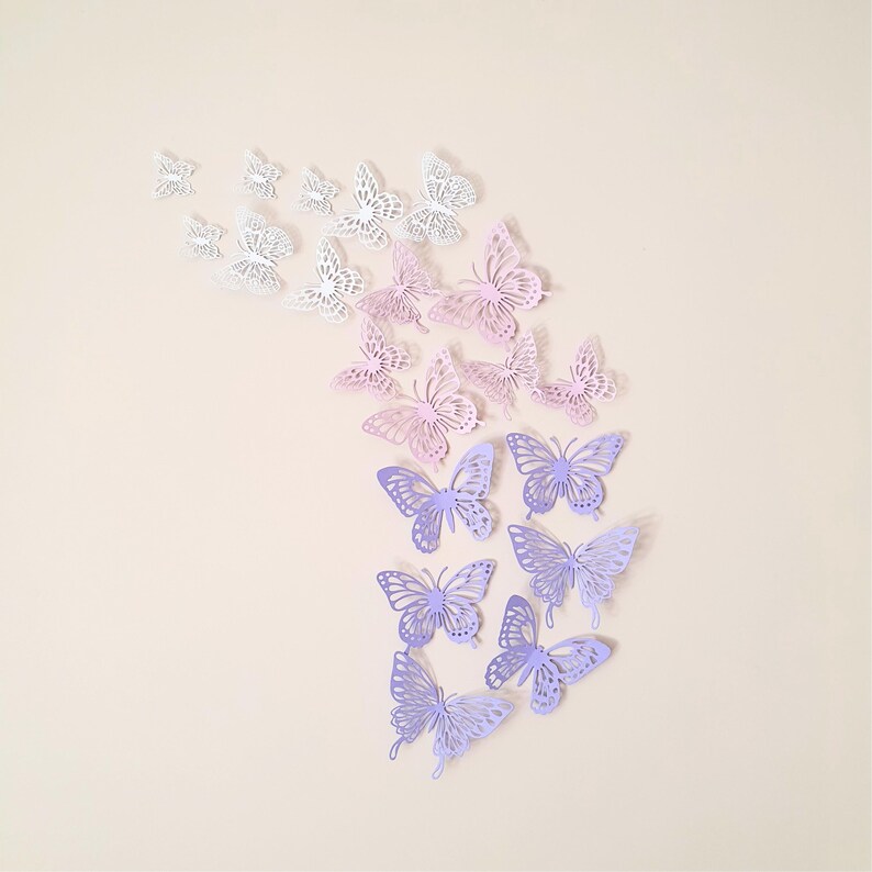 May include: A collection of paper butterflies in various sizes and shades of white, pink, and purple. The butterflies are arranged in a cascading pattern, creating a decorative wall art piece. The butterflies have intricate cut-out designs.