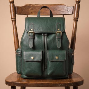 Puede incluir: Una mochila de cuero verde oscuro con una solapa superior con dos correas de hebilla, un cierre de cordón y dos bolsillos delanteros con botones a presión. La bolsa incluye compartimentos laterales con cremallera y un asa superior texturizada.