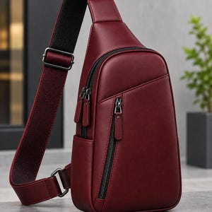 Puede incluir: Un bolso bandolera de color burdeos intenso con un acabado mate liso, una correa de hombro ajustable única con forro negro texturizado y dos compartimentos frontales con cremallera, herrajes de metal oscuro y tiradores a juego.