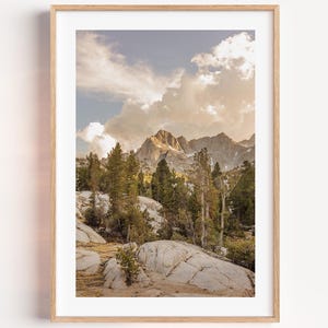May include: A framed landscape art print featuring a mountain scene. The artwork showcases a mountain range under a cloudy sky with golden hues. The foreground displays rocks and trees, creating a natural, scenic view. The print is encased in a light-colored wooden frame, suitable for home decor.