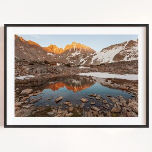 May include: A framed landscape photograph featuring a mountain range with snow-capped peaks and a reflective lake. The mountains are illuminated by the setting sun, casting warm orange hues. The foreground is rocky, with a clear sky above.