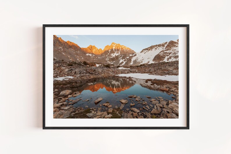 May include: A framed landscape photograph featuring a mountain range with snow-capped peaks and a reflective lake. The mountains are illuminated by the setting sun, casting warm orange hues. The foreground is rocky, with a clear sky above.