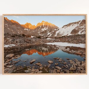 May include: A framed landscape print featuring a mountain range with snow-capped peaks and a reflective lake. The mountains are illuminated by the golden light of the setting sun, casting a warm glow on the scene. The water reflects the mountains.