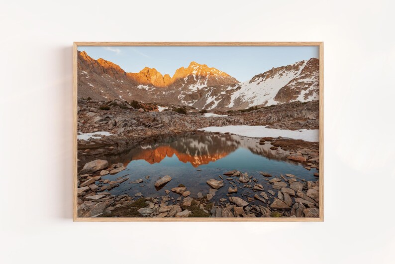 May include: A framed landscape print featuring a mountain range with snow-capped peaks and a reflective lake. The mountains are illuminated by the golden light of the setting sun, casting a warm glow on the scene. The water reflects the mountains.