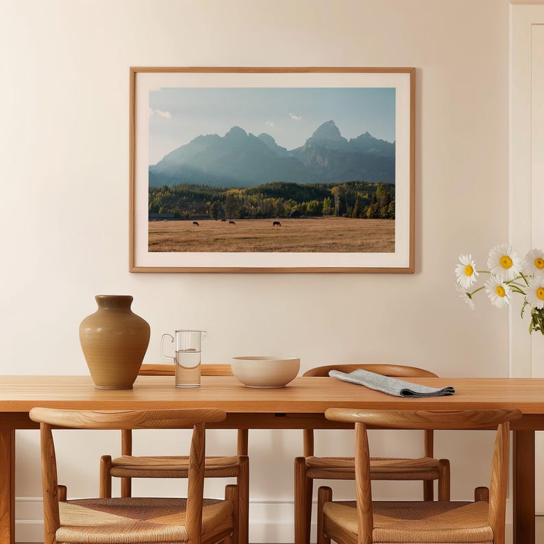 May include: A framed landscape print featuring a mountain range under a blue sky. The print is displayed above a wooden dining table with chairs. A vase, pitcher, and bowl are on the table. Daisies in a vase are on the right.