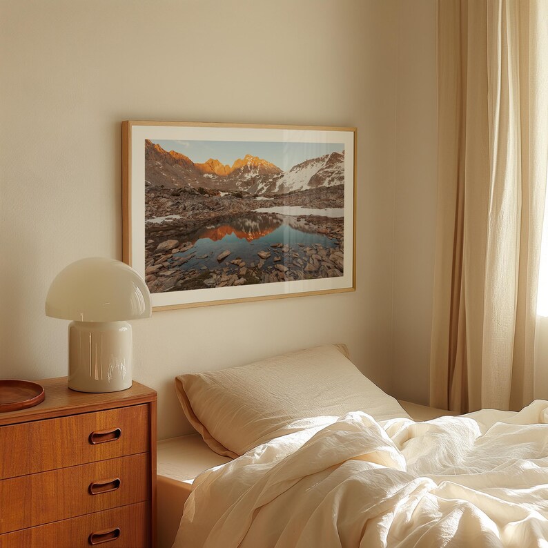 May include: A framed landscape photograph depicts a mountain range reflected in a lake. The image features a natural color palette with shades of brown, blue, and white. The photograph is displayed above a bed with cream-colored linens.