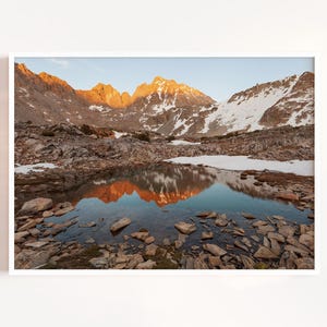 May include: A framed landscape photograph captures a mountain range at sunrise. The peaks are illuminated with golden light, reflected in a still pool of water. The foreground is filled with rocks and the sky is a soft blue.